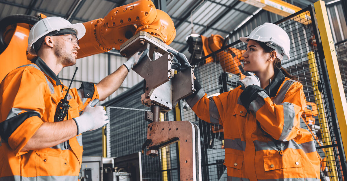 Two engineers in hard hats and orange jackets service a robot welding machine in an automated factory.