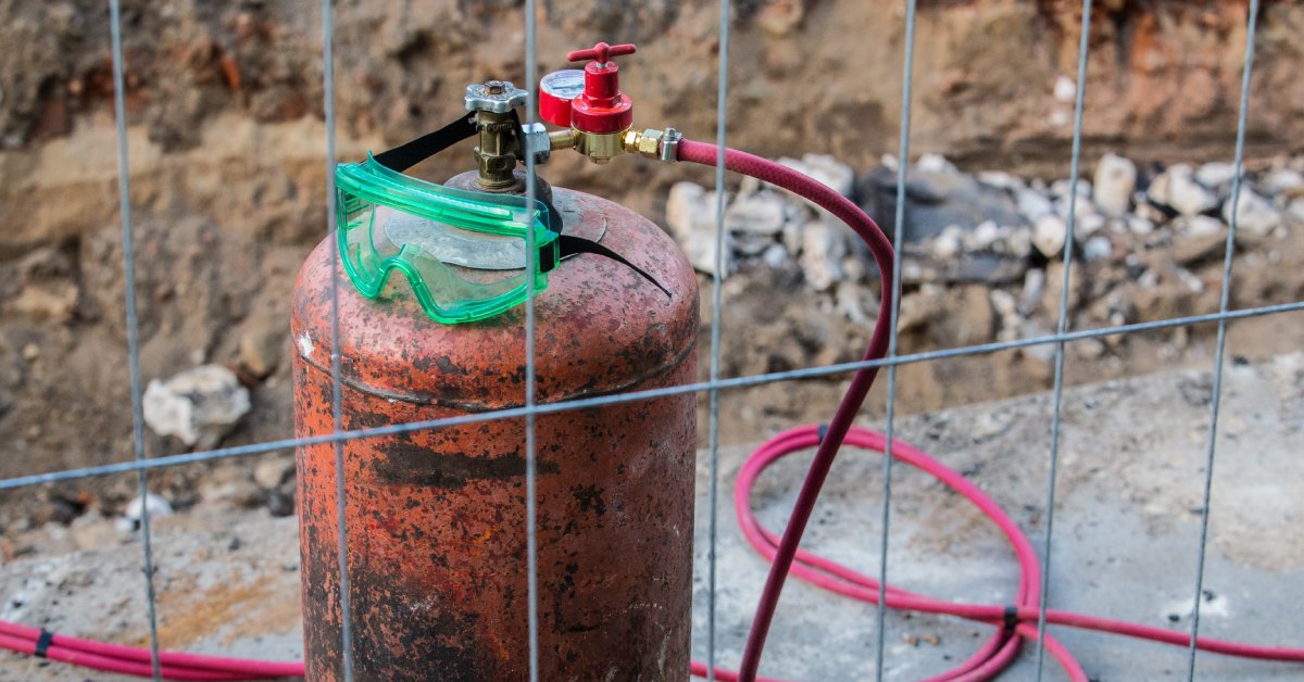 A pair of protective goggles rests on a gas tank in a dug-out site. A fence separates the equipment.