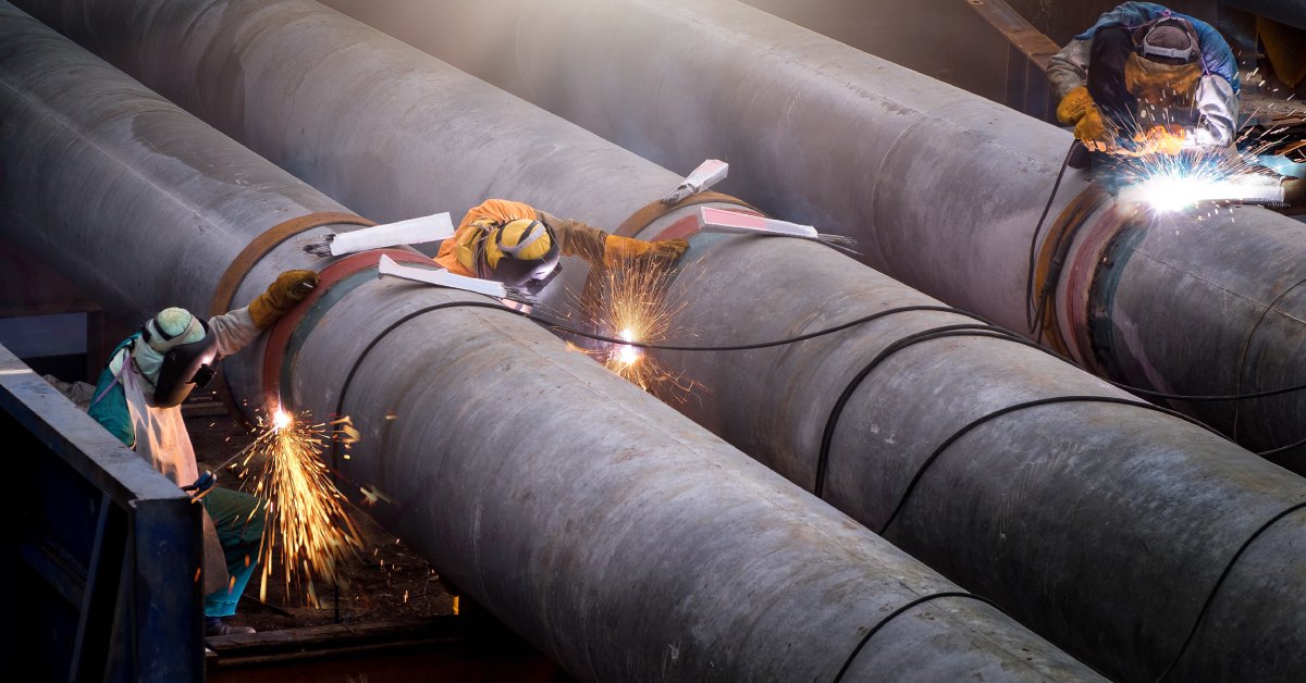 Three welders are assembling three large pipes at a construction site using professional equipment and safety gear.