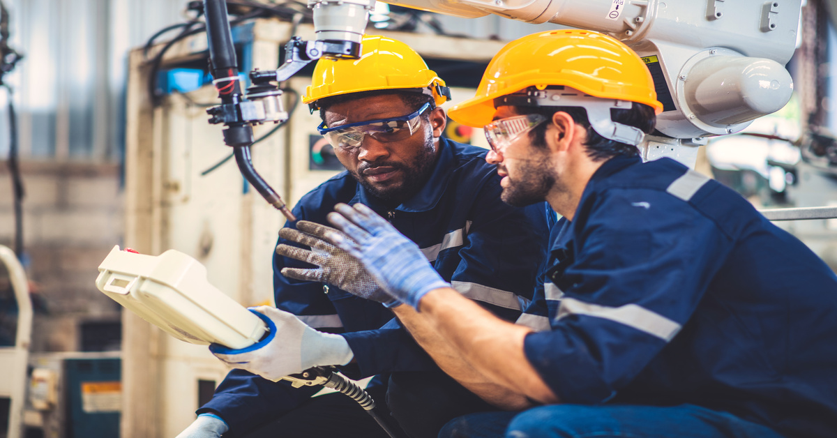 Two men in safety glasses and hard hats stand beneath a robotic arm with a welding torch as one holds a controller.
