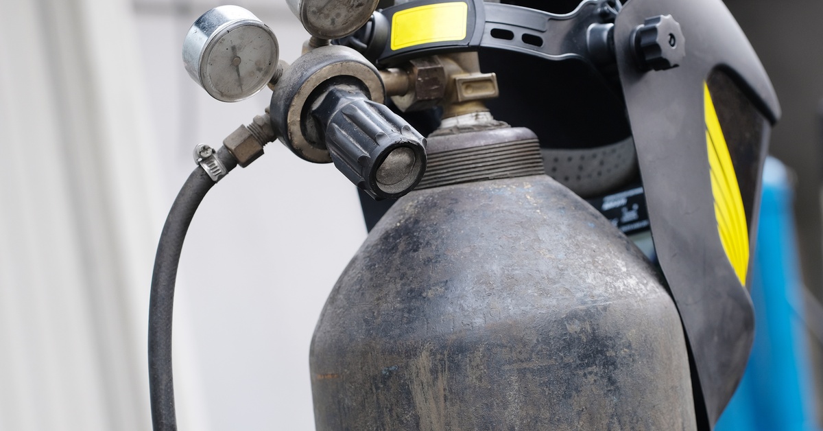 A black welding helmet with yellow detailing in the front, resting on top of a worn, unlabeled black gas cylinder.